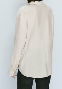 Chemise à manches longues en tissu beige clair, dotée d'un col à revers, d'une coupe décontractée et d'un ourlet arrondi. Vue de dos montrant un design simple et des poignets à boutons.