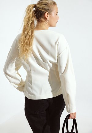 Baltas ilgomis rankovėmis palaidinė su struktūruotu dizainu, turinti laisvą siluetą ir subtilią tekstūrą, derinama su juodomis kelnėmis ir maža juoda rankine.