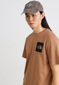 Camiseta de algodón marrón con un parche de logo negro en el lado izquierdo del pecho, combinada con una gorra gris que muestra un diseño gráfico blanco.