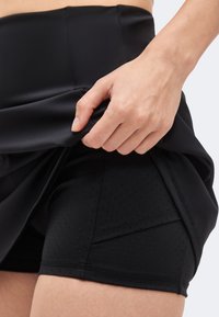 Main soulevant une jupe de sport noire pour révéler un short noir ajusté perforé avec une poche latérale sur la cuisse.