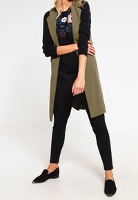 Chaleco sin mangas de color verde oliva sobre un suéter negro con bordados florales, combinado con pantalones ajustados negros y zapatos negros de tipo slip-on.