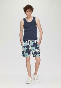 Marineblauw tanktop, gestreepte zwemshorts met grote lichtblauwe en crèmekleurige bloemenpatronen, en witte sneakers. Casual, relaxte pasvorm.