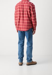 Czerwona i beżowa koszula flanelowa w kratę, noszona z niebieskimi dżinsami i brązowymi butami. Koszula ma zaokrąglony dół i mankiety zapinane na guziki.