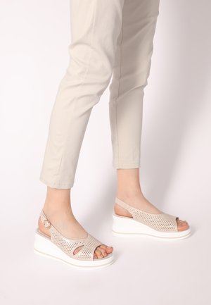 Beige Keilsandaletten mit durchlöchertem Oberdesign und verstellbarem Knöchelriemen. Weiße Plattformsohle. Fuß teilweise sichtbar.