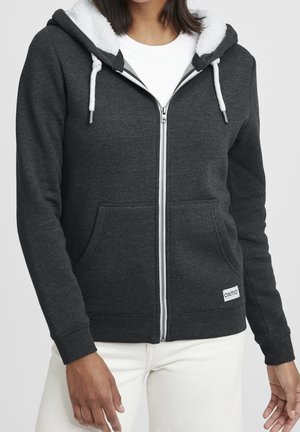 Femme portant un sweat à capuche zippé gris foncé avec doublure en polaire blanche et cordons, associée à un pantalon blanc, debout devant un fond uni.