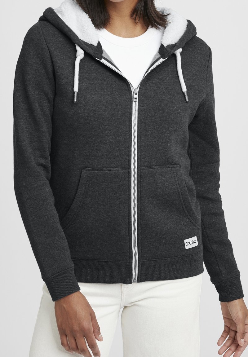 Femme portant un sweat à capuche zippé gris foncé avec doublure en polaire blanche et cordons, associée à un pantalon blanc, debout devant un fond uni.