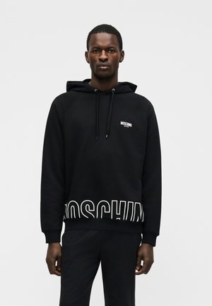 Homme portant un sweat à capuche Moschino noir et un pantalon assorti, debout devant un fond gris clair uni, face à l'avant avec une expression neutre.