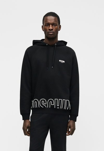 Άνδρας με μαύρο φούτερ Moschino με κουκούλα και αντίστοιχο παντελόνι, στέκεται μπροστά σε απλό ανοιχτό γκρι φόντο, κοιτάζοντας μπροστά με ουδέτερη έκφραση.