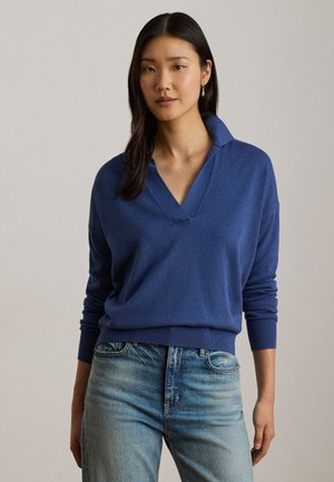 Kvinde med langt mørkt hår iført en blå V-hals sweater, lyseblå jeans og en guldring ørepynt, stående mod en ensfarvet baggrund.