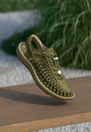 Olijfgroene geweven sandaal met verstelbare bungee-veters zwevend boven een houten oppervlak tegen een vervaagde groene achtergrond.