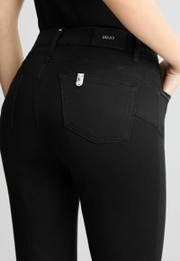 Jean noir à texture lisse, avec une étiquette LIU-JO, une quincaillerie argentée sur la poche arrière, et une coupe ajustée.
