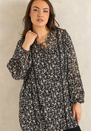 Kobieta ubrana w czarną, długą sukienkę w kwiaty z przezroczystymi rękawami i wiązaniem przy szyi, pozująca na jasnym, jednolitym tle.