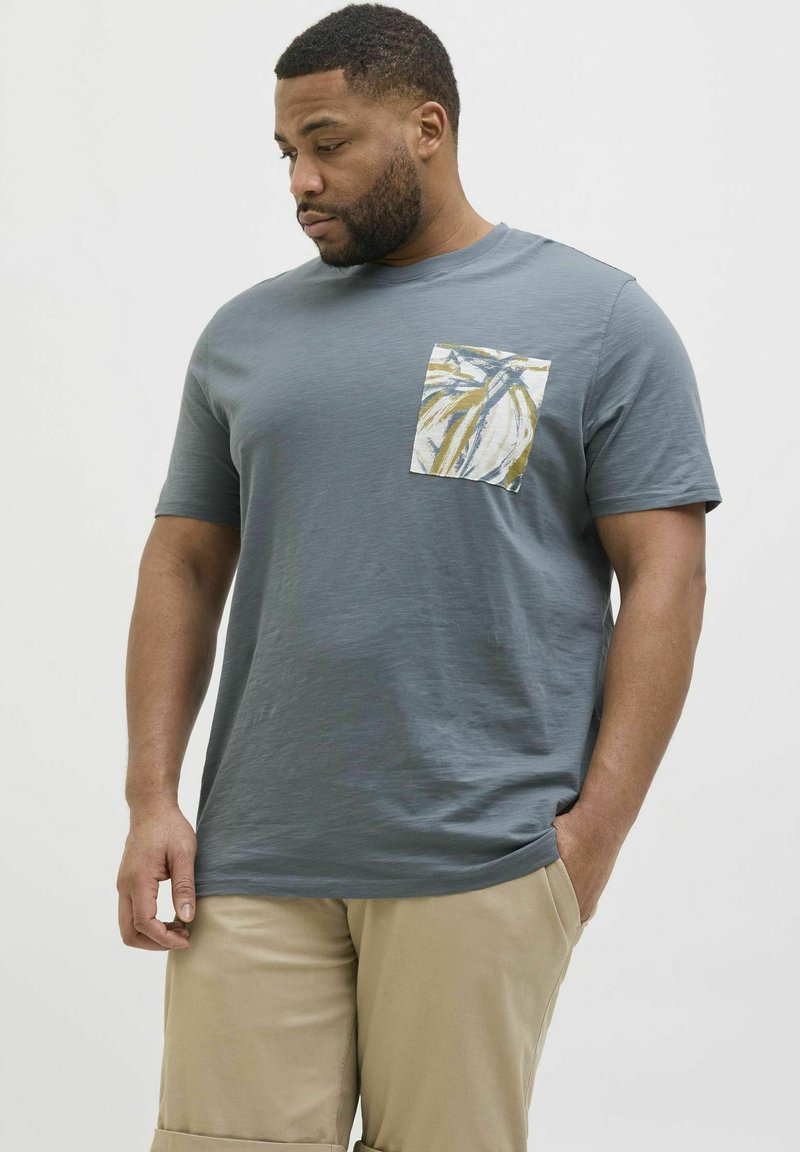 T-shirt gris à manches courtes avec une poche à motifs comportant des feuilles jaunes et bleues. Le tissu semble doux avec une coupe décontractée.