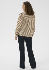 Donna con i capelli ricci che indossa una blusa a righe verticali marrone e bianco e jeans blu scuro, in piedi con le spalle rivolte verso la fotocamera.