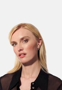 Blont hår, ljus hy, bär en svart transparent skjorta med knappar. Notabla drag: subtil sminkning, hoop örhängen och neutral läppfärg.