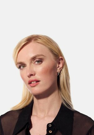 Blont hår, ljus hy, bär en svart transparent skjorta med knappar. Notabla drag: subtil sminkning, hoop örhängen och neutral läppfärg.