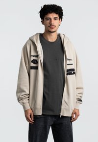 Beige zip-up hættejakke med broderede mærker, der har en rummelig pasform og ribbede cuffs, lagdelt over en mørkegrå t-shirt og parret med mørke jeans.