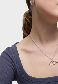 Collar de plata con un charm de corazón entrelazado y una flecha en una delicada cadena, llevado con un top gris acanalado que resalta el escote.