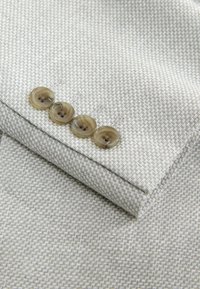 Manche de veste gris clair avec quatre boutons bruns translucides et un motif texturé subtil.