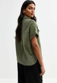 Groene kortebloushirt, geweven stof, relaxed fit, met een kraag en zakken. Model toont de achterkant tegen een neutrale achtergrond.