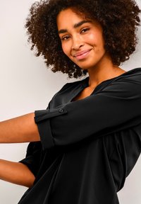 Blouse noire ample avec un col rond, des poignets boutonnés et un tissu doux. Présente des drapés fluides aux épaules et aux manches.