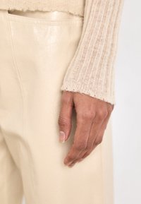 Beige svetr s žebrovanými rukávy a třepeními, spárovaný s hladkými, světlými koženými kalhotami. Ruka jemně spočívá na noze.