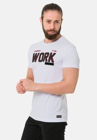 Weißes Baumwoll-T-Shirt mit dem Wort "WORK" in fetter, strukturiertem schwarzen Schriftzug. Kurze Ärmel, runder Halsausschnitt und schwarze Akzente am Saum.