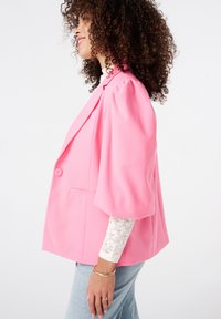 Roze, oversized blazer met pofmouwen, enkele knoopsluiting, zijzakken, gladde stof, gedragen over een witte kanten top en jeans.