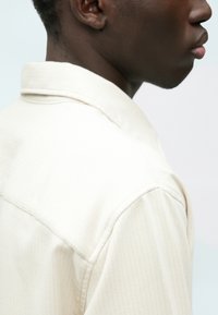 Chemise col beige clair avec de subtils rayures verticales, présentant une coupe décontractée et une texture douce, vue de côté.