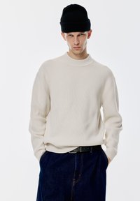 Rippiges cremefarbenes Sweatshirt mit hohem Kragen und langen Ärmeln, kombiniert mit einer schwarzen Strickmütze und dunklen Jeans.