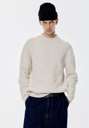 Rippiges cremefarbenes Sweatshirt mit hohem Kragen und langen Ärmeln, kombiniert mit einer schwarzen Strickmütze und dunklen Jeans.