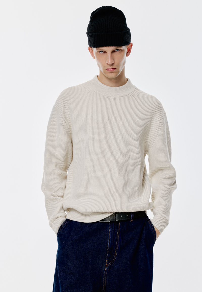 Rippiges cremefarbenes Sweatshirt mit hohem Kragen und langen Ärmeln, kombiniert mit einer schwarzen Strickmütze und dunklen Jeans.
