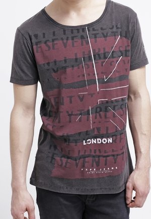 Mann trägt ein dunkelgraues T-Shirt mit einem in Burgunderrot und Schwarz gehaltenen abstrakten Muster und dem Schriftzug "London" auf der Vorderseite.