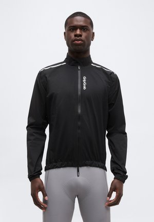 Homme portant une veste noire GripGrab zippée et un pantalon de cyclisme rembourré gris, debout face à l'avant sur un fond uni.