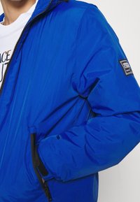 Blå puffjacka med dragkedja, sidofickor och en patch på ärmen. Tillverkad av lätt, texturerat tyg.