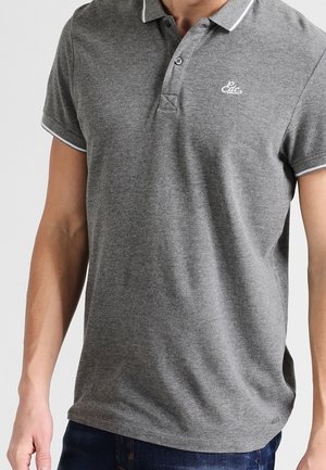 Homme portant un polo gris à manches courtes avec bordure blanche et petit logo sur la poitrine gauche, associé à un jean bleu foncé.