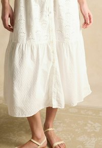 Weißes Baumwollkleid mit Lochstickerei, Knopfriegelung und gestuftem Rock. Getragen mit hellen Sandalen.