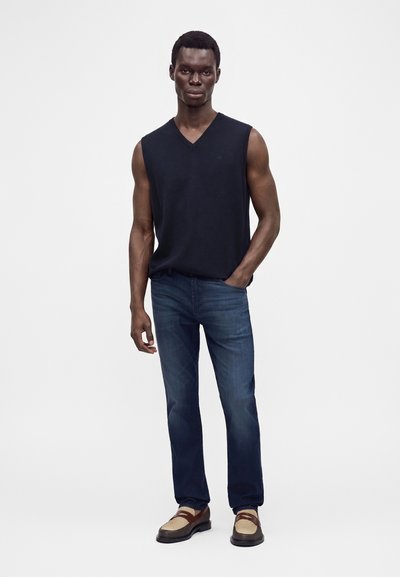 Homme portant un t-shirt noir sans manches à col en V, un jean bleu et des mocassins marron bicolores, debout avec une main dans la poche.