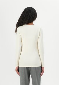 Langärmliges Shirt aus weichem cremefarbenem Stoff, mit runder Halslinie und taillierter Passform. Kombiniert mit grauen Hosen, die eine glatte Textur aufweisen.