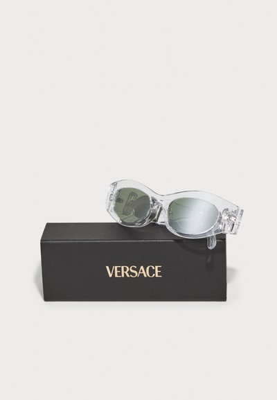 Ochelari de soare din acetat transparent cu lentile verzi, așezate pe o cutie neagră. Cutia prezintă logo-ul auriu "VERSACE" afisat proeminent.