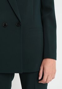 Ciemnozielony, dwurzędowy żakiet wykonany z fakturowanej tkaniny. Posiada czarne guziki oraz jedną boczną kieszeń. Dopasowany krój, gładkie rękawy.