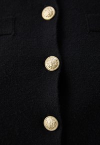 Black cardigan featuring three gold buttons with intricate designs, textured fabric, and two small pockets on the front.