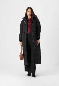 Sort puffet jakke med et langt, quiltet design, der har en frontlynlås og sidelommer. Båret over en bordeaux bluse og sorte bukser.