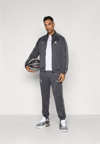 Grå træningsdragt med hvide detaljer, der inkluderer en lynlåsjakke og elastiske bukser. Tilbehør: sort basketball med logo. Fodtøj: grå sneakers.