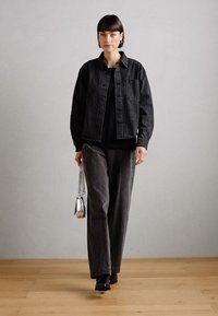 Chaqueta de mezclilla negra con bolsillos frontales, combinada con pantalones sueltos de un gris oscuro. Los accesorios incluyen un pequeño bolso de mano plateado. Fondo simple.