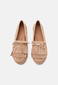 Μπεζ μοκασίνια από σουέτ με στρωματοποιημένες λεπτομέρειες φράντζας και δεμένους φιόγκους στην κορυφή, εμφανιζόμενα από πάνω σε λευκό φόντο.