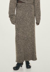 Lange, gebreide rok in een zacht bruin en crèmekleurig patroon; heeft een aansluitende taille, lange mouwen en een geribbelde zoom. Gecombineerd met zwarte klompen.