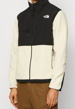 Mężczyzna noszący czarno-kremową polarową kurtkę North Face zapinaną na suwak, z zapinanymi na suwak kieszeniami na piersi i bokach, w połączeniu z brązowymi spodniami.