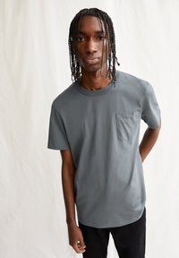 Grijze katoenen t-shirt met een ronde hals en een borstzak, gedragen door een persoon met vlechten, tegen een neutrale achtergrond.