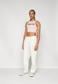 Cropped, multicolored striped tank top with text "PROUD TO BE" paired with loose-fitting white trousers and gray sneakers.